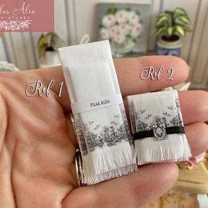 Puede incluir: Dos toallas de mano blancas con dise&ntilde;os florales en blanco y negro y ribete de flecos. Las toallas est&aacute;n etiquetadas como "Ref 1" y "Ref 2".