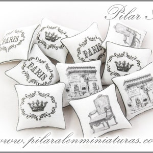 May include: A collection of nine white miniature pillows with black and gray embroidery. The pillows feature various designs, including the word "Paris", a crown, and an image of the Arc de Triomphe. The pillows are arranged on a white surface. www.pilaralenminiaturas.com