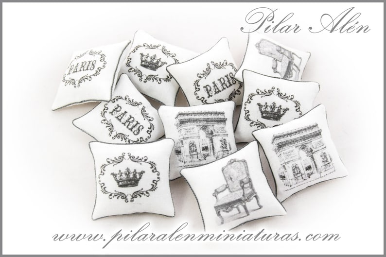 May include: A collection of nine white miniature pillows with black and gray embroidery. The pillows feature various designs, including the word "Paris", a crown, and an image of the Arc de Triomphe. The pillows are arranged on a white surface. www.pilaralenminiaturas.com