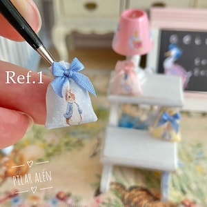 K&ouml;nnte beinhalten: Eine kleine blaue Stofftasche mit einer blauen Schleife und einem Peter Rabbit-Druck. Die Tasche wird von einer Hand gehalten. Ref.1 ist auf dem Bild geschrieben.