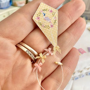 May include: A miniature kite with a yellow gingham pattern and a floral and bird design. The kite has pink and yellow bows attached to the string. The kite is a craft item.