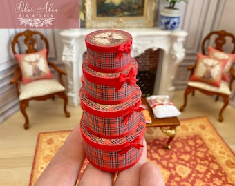 May include: A stack of four miniature round gift boxes featuring a red and black plaid pattern. Each box is wrapped with a thin red ribbon tied into a small bow on the front. The top box lid displays a circular illustration of a deer head.