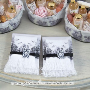Puede incluir: Dos toallas de mano blancas con dise&ntilde;os impresos en blanco y negro y bordes con flecos. Las toallas est&aacute;n sobre una superficie de madera clara.