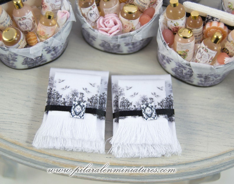 Puede incluir: Dos toallas de mano blancas con dise&ntilde;os impresos en blanco y negro y bordes con flecos. Las toallas est&aacute;n sobre una superficie de madera clara.
