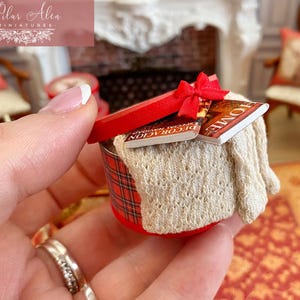 May include: Miniature red and plaid round box with a red lid tied with a bow. The box contains a cream-colored knitted blanket and two miniature books. The books have the word "HOME" on the cover.