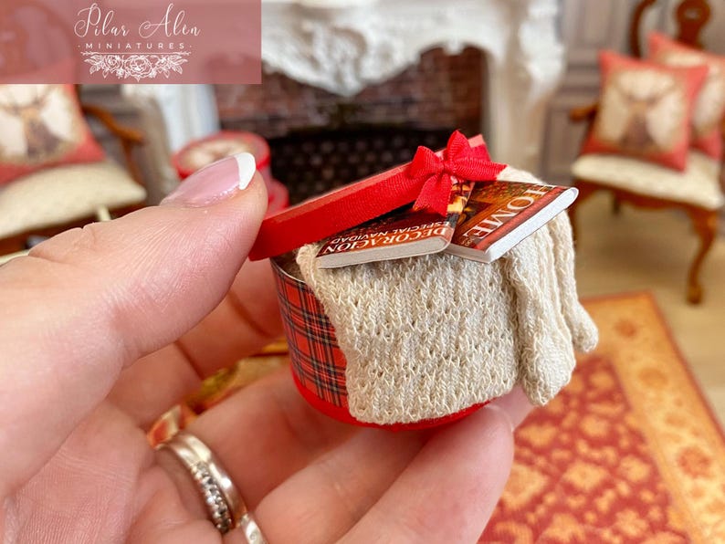 May include: Miniature red and plaid round box with a red lid tied with a bow. The box contains a cream-colored knitted blanket and two miniature books. The books have the word "HOME" on the cover.