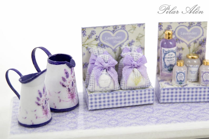 May include: A miniature display featuring two white pitchers with purple lavender patterns, four small fabric sachets with lavender bows, and a set of miniature bottles with gold caps, all arranged on a lavender patterned base with the text Pilar Alen.