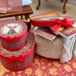 May include: Miniature red plaid gift boxes with red bows, stacked on a red patterned surface. The top box features a deer illustration. A small, off-white knitted blanket holds two miniature books, one with the word "HOME" on the cover.