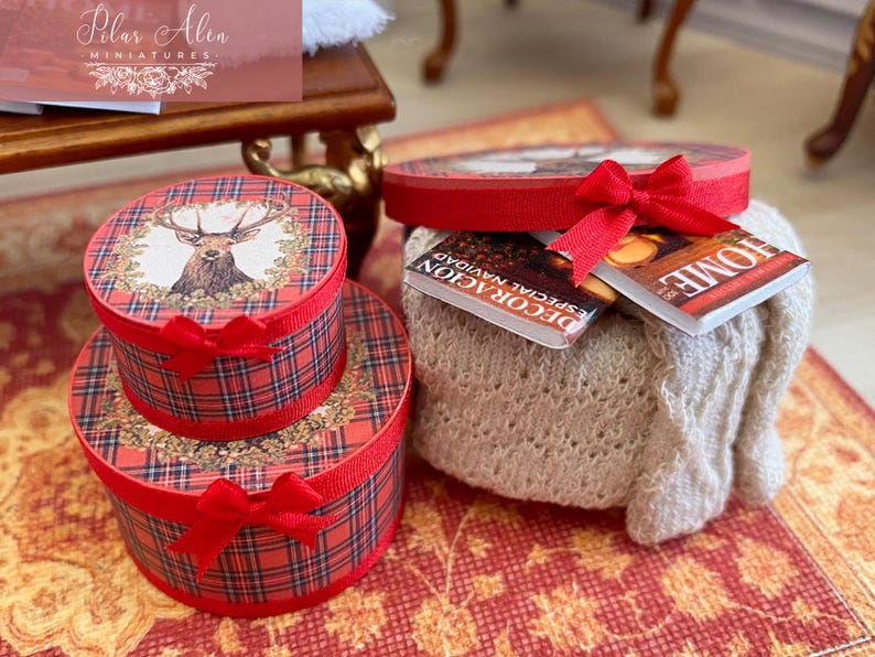 May include: Miniature red plaid gift boxes with red bows, stacked on a red patterned surface. The top box features a deer illustration. A small, off-white knitted blanket holds two miniature books, one with the word "HOME" on the cover.