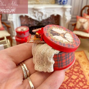 May include: A miniature round box with a red plaid pattern and a deer illustration on the lid. The box contains a small, cream-colored knitted item. A miniature book with the word "HOME" is visible.