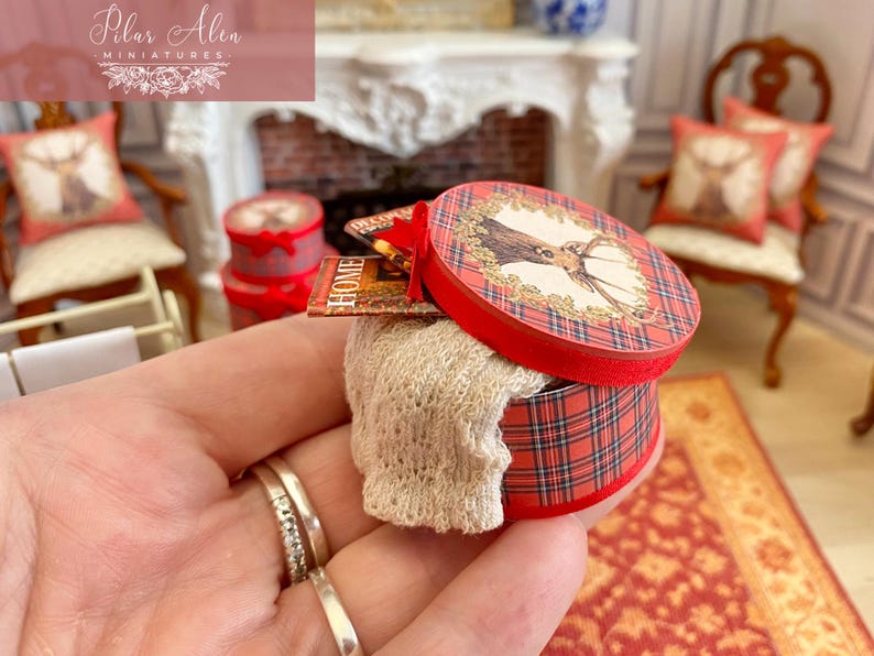 May include: A miniature round box with a red plaid pattern and a deer illustration on the lid. The box contains a small, cream-colored knitted item. A miniature book with the word "HOME" is visible.