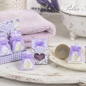 May include: Miniature lavender sachets in a purple gingham display box and individual packaging. Each sachet features a lavender sprig print and a purple ribbon bow. A one-euro coin is placed next to a sachet for scale, showing a height of 1.5 cm.