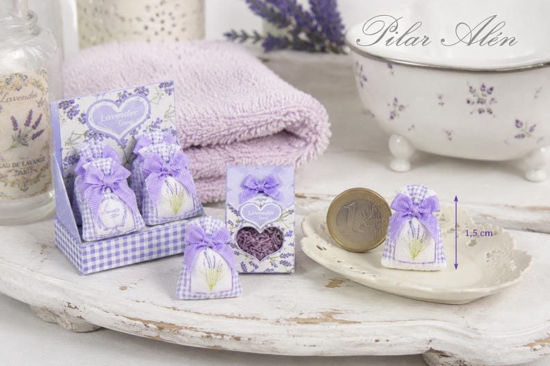 May include: Miniature lavender sachets in a purple gingham display box and individual packaging. Each sachet features a lavender sprig print and a purple ribbon bow. A one-euro coin is placed next to a sachet for scale, showing a height of 1.5 cm.