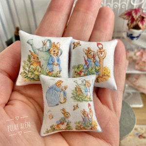 Può includere: Tre cuscini in miniatura con sfondo bianco e un motivo ripetuto di personaggi di Peter Rabbit. I cuscini sono in tessuto e hanno un bordo blu chiaro.