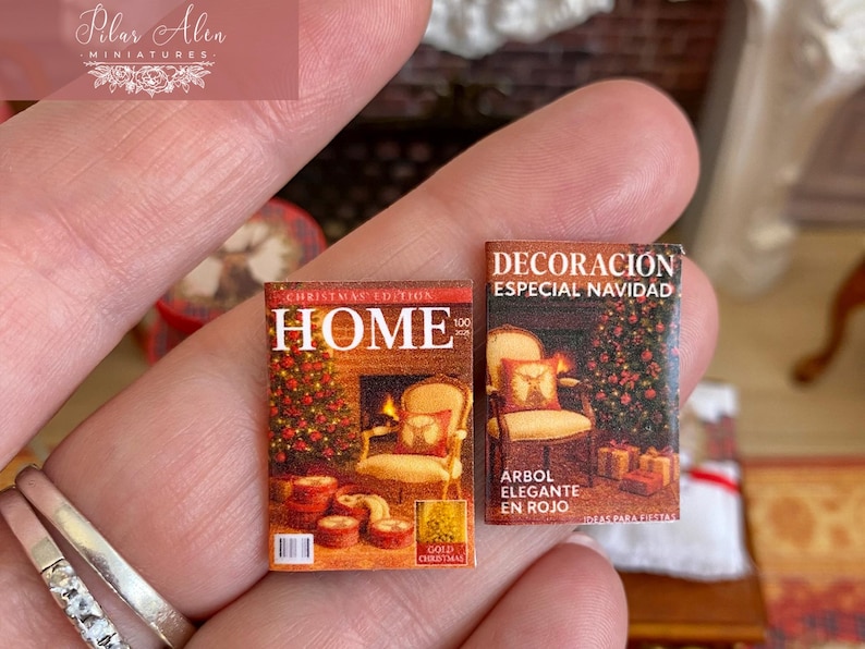 May include: Miniature Christmas magazines with detailed covers. One features a fireplace and the word "HOME", the other says "DECORACION ESPECIAL NAVIDAD". Both are held in a hand, showcasing their small size.