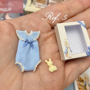 Könnte beinhalten: Miniatur-Baby-Kleidungsset. Ein hellblauer Strampler mit weißer Bordüre und Schleife, eine kleine gelbe Hasenfigur und eine kleine Schachtel mit einer Peter-Rabbit-Illustration und einer blauen Schleife.