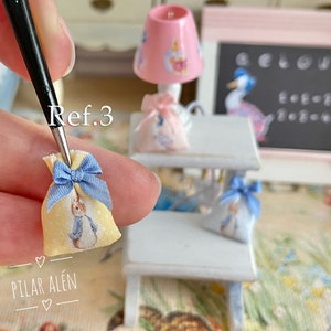 K&ouml;nnte beinhalten: Eine kleine, gelbe Stofftasche mit einer blauen Schleife und einem Peter-Rabbit-Motiv. Die Tasche wird von einer Hand mit einer Pinzette gehalten. Der Text "Ref. 3" ist auf dem Bild zu sehen. Der Text "PILAR AL&Eacute;N" ist ebenfalls auf dem Bild zu sehen.