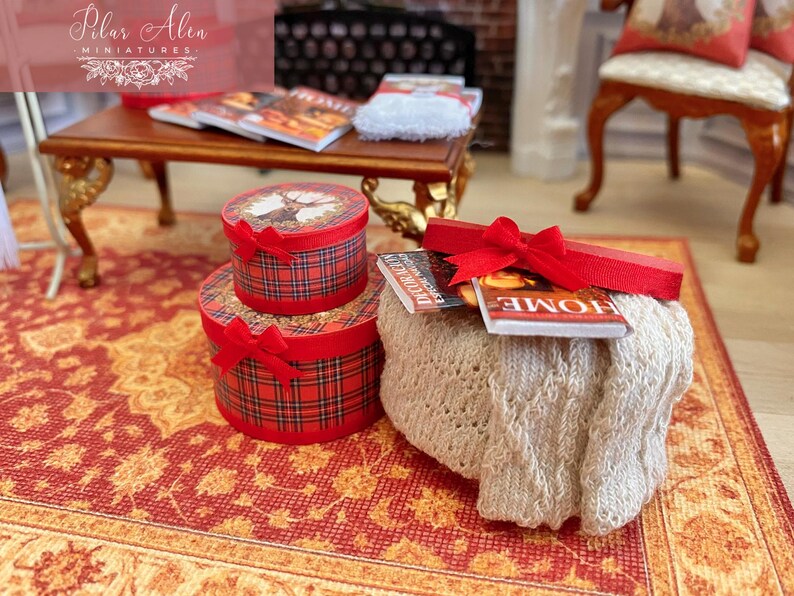 May include: Miniature Christmas-themed decor featuring two plaid gift boxes with red bows, a small table with magazines, and a decorative knitted pouf with more magazines and a red ribbon. The scene is set on a patterned rug.