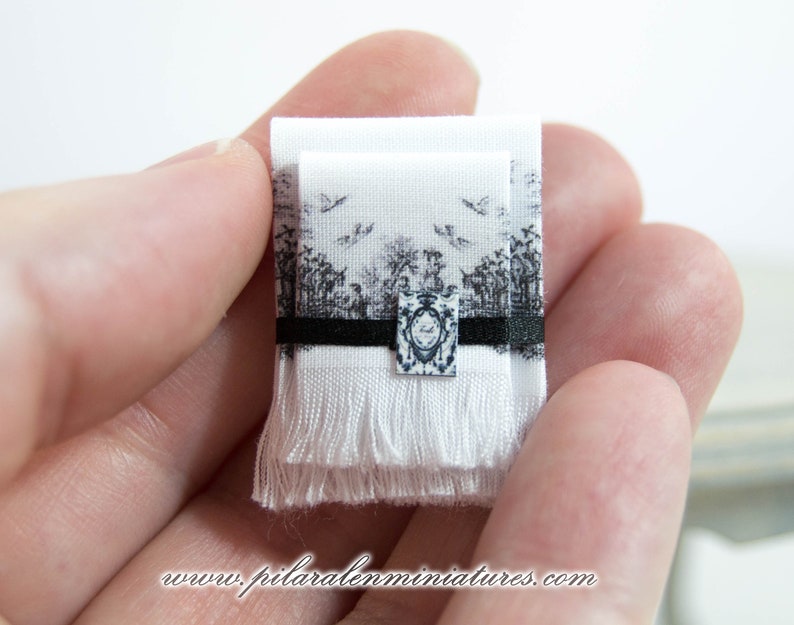 Puede incluir: Una toalla de tela blanca de tama&ntilde;o para casa de mu&ntilde;ecas con un borde negro y un dise&ntilde;o impreso en blanco y negro. La toalla tiene un fleco en la parte inferior.