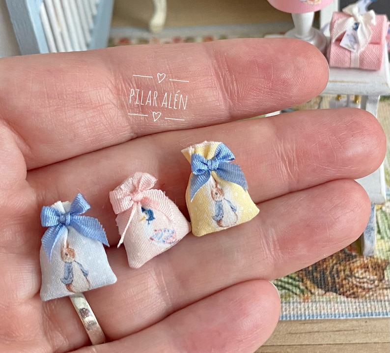 K&ouml;nnte beinhalten: Drei kleine Stoffgeschenkt&uuml;ten mit blauem, rosa und gelbem Stoff. Jede Tasche hat ein blaues Band und ein Peter-Rabbit-Design. Die Taschen werden in einer Hand gehalten.