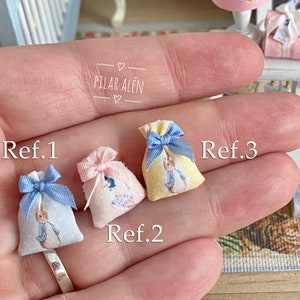 K&ouml;nnte beinhalten: Drei kleine Stoffbeutel mit Peter Rabbit-Motiven. Die Beutel sind in Blau, Rosa und Gelb mit blauen Schleifenb&auml;ndern. Die Beutel sind mit Ref. 1, Ref. 2 und Ref. 3 beschriftet.