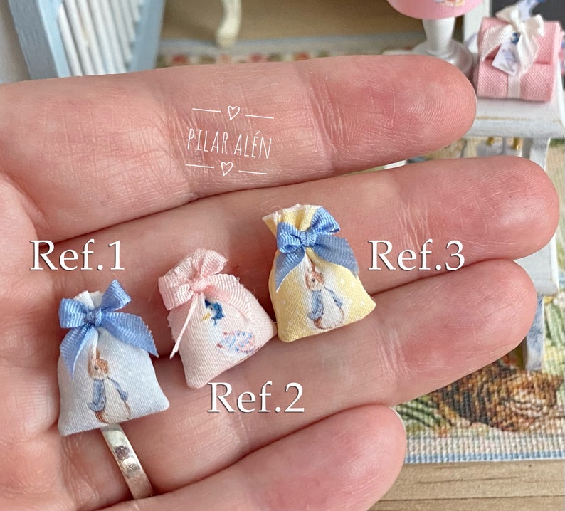 K&ouml;nnte beinhalten: Drei kleine Stoffbeutel mit Peter Rabbit-Motiven. Die Beutel sind in Blau, Rosa und Gelb mit blauen Schleifenb&auml;ndern. Die Beutel sind mit Ref. 1, Ref. 2 und Ref. 3 beschriftet.