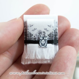 Puede incluir: Una toalla de tela blanca de tama&ntilde;o para casa de mu&ntilde;ecas con un borde negro y un dise&ntilde;o impreso en blanco y negro. La toalla tiene un fleco en la parte inferior.