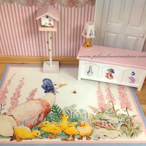 May include: Miniature dollhouse scene featuring a rug with a duck and ducklings illustration, a white and pink chest with bunny illustrations, a birdhouse, and a lamp. The scene is set against a pink striped wall.