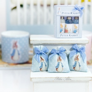 K&ouml;nnte beinhalten: Drei blaue und wei&szlig;e Stoffbeutel mit Peter Rabbit-Motiven und blauen Schleifen, die auf einem wei&szlig;en Regal mit einem Peter Rabbit-Geschenkset mit drei Flaschen blauer Fl&uuml;ssigkeit pr&auml;sentiert werden. Das Regal hat ein wei&szlig;es Schild mit der Aufschrift "Pilar Allen".