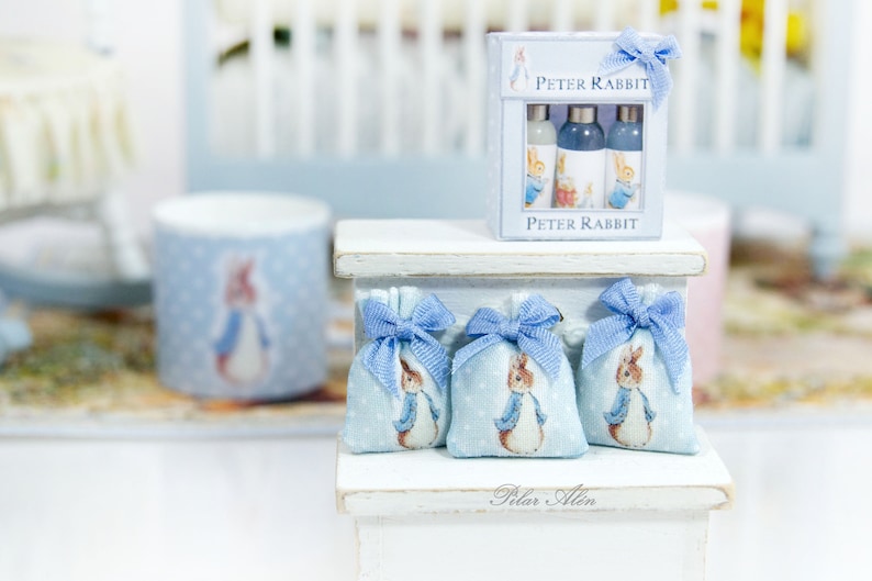 K&ouml;nnte beinhalten: Drei blaue und wei&szlig;e Stoffbeutel mit Peter Rabbit-Motiven und blauen Schleifen, die auf einem wei&szlig;en Regal mit einem Peter Rabbit-Geschenkset mit drei Flaschen blauer Fl&uuml;ssigkeit pr&auml;sentiert werden. Das Regal hat ein wei&szlig;es Schild mit der Aufschrift "Pilar Allen".