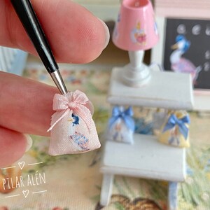 K&ouml;nnte beinhalten: Eine kleine rosa Stofftasche mit einem blauen und wei&szlig;en Blumenmuster und einem rosa Band. Die Tasche wird von einer Hand gehalten. Die Tasche liegt auf einem wei&szlig;en Regal mit zwei weiteren kleinen Taschen.