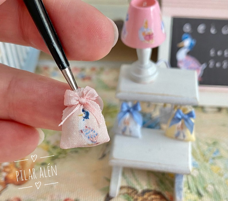 K&ouml;nnte beinhalten: Eine kleine rosa Stofftasche mit einem blauen und wei&szlig;en Blumenmuster und einem rosa Band. Die Tasche wird von einer Hand gehalten. Die Tasche liegt auf einem wei&szlig;en Regal mit zwei weiteren kleinen Taschen.