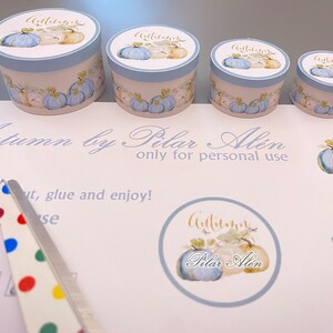 May include: Four round blue and white gift boxes with a watercolor design of pumpkins and leaves. The boxes are labeled "Autumn" in gold lettering. The boxes are on a white surface with a sheet of paper that says "Autumn by Pilar Alen only for personal use".