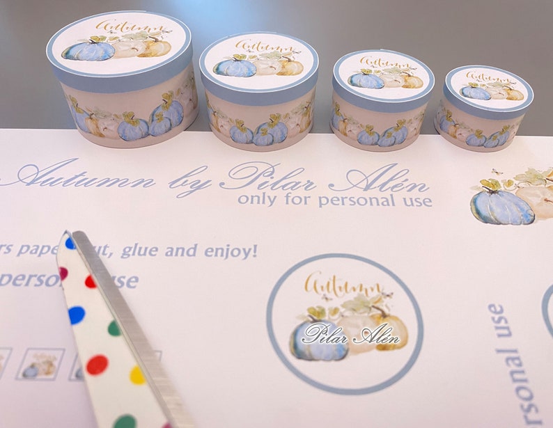 May include: Four round blue and white gift boxes with a watercolor design of pumpkins and leaves. The boxes are labeled "Autumn" in gold lettering. The boxes are on a white surface with a sheet of paper that says "Autumn by Pilar Alen only for personal use".