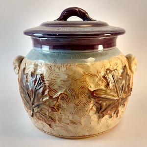 Puede incluir: Una olla de frijoles de cerámica con tapa, decorada con un diseño de hoja de arce. La olla está esmaltada en tonos de marrón, beige y azul. La tapa tiene un esmalte morado. La olla está etiquetada "Maple Leaf Bean Pot No. 601".