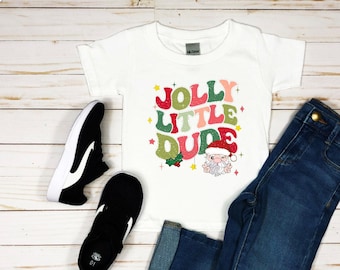 May include: White t-shirt with a colorful retro design that says "Jolly Little Dude" with a cartoon Santa Claus wearing a red hat. The shirt is paired with blue jeans and black sneakers.