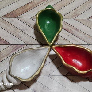 May include: Three decorative seashell-shaped bowls, one green, one white, and one red, with gold accents. The bowls are arranged in a triangular pattern on a wooden surface.