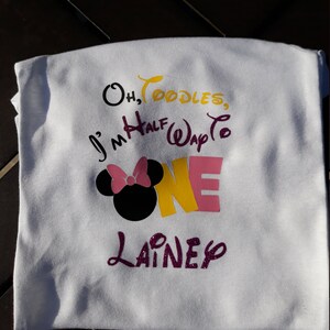 May include: White t-shirt with colorful text and graphics. The shirt features the phrase "Oh, Toodles, I'm Half Way to ONE" with a Minnie Mouse head and the name "Lainey". The text is in black, yellow, pink, and purple.
