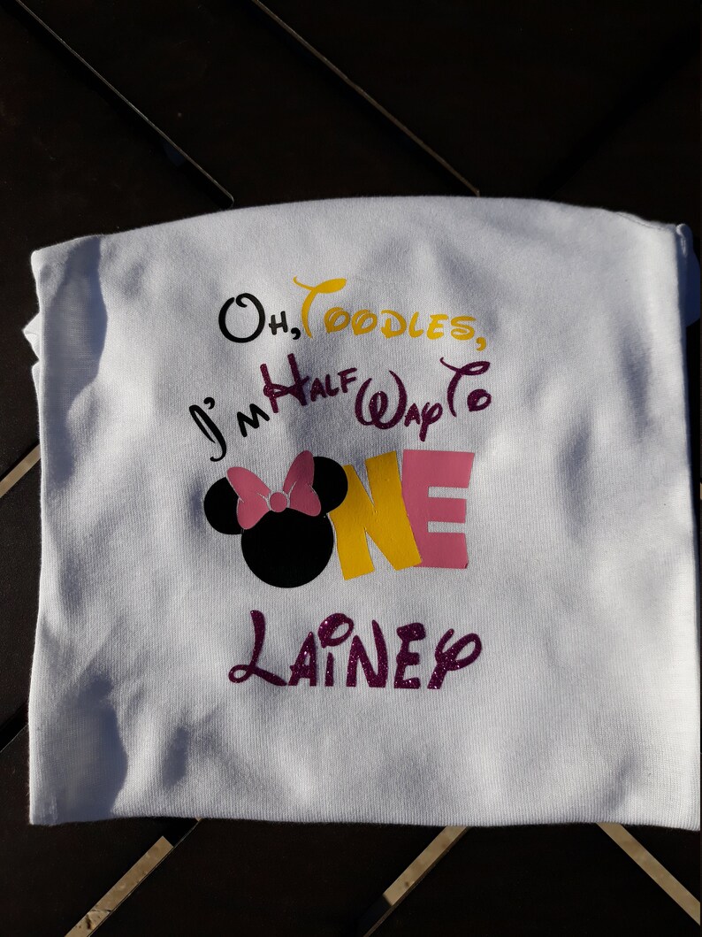 May include: White t-shirt with colorful text and graphics. The shirt features the phrase "Oh, Toodles, I'm Half Way to ONE" with a Minnie Mouse head and the name "Lainey". The text is in black, yellow, pink, and purple.