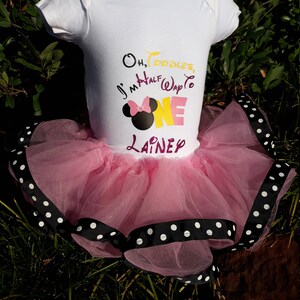 May include: A white baby bodysuit with the text "Oh, Toodles, I'm Half Way to ONE Lainey" and a Minnie Mouse graphic. The bodysuit is paired with a pink tulle tutu trimmed with black ribbon and white polka dots.