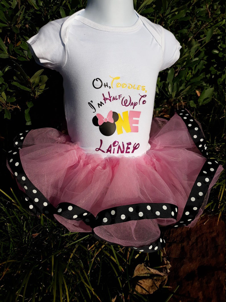 May include: A white baby bodysuit with the text "Oh, Toodles, I'm Half Way to ONE Lainey" and a Minnie Mouse graphic. The bodysuit is paired with a pink tulle tutu trimmed with black ribbon and white polka dots.