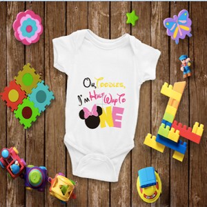 May include: White baby onesie with the text "Oh, Toodles, I'm Half Way to ONE" and a Minnie Mouse graphic. The text is in black, yellow, pink, and white. The onesie is surrounded by colorful toys on a wooden background.