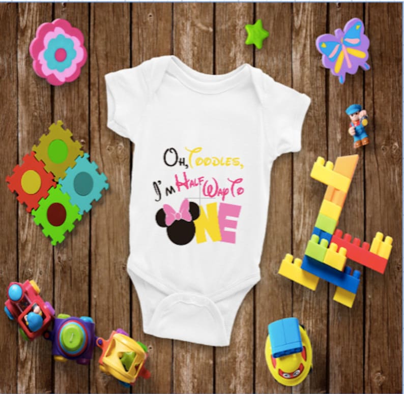 May include: White baby onesie with the text "Oh, Toodles, I'm Half Way to ONE" and a Minnie Mouse graphic. The text is in black, yellow, pink, and white. The onesie is surrounded by colorful toys on a wooden background.