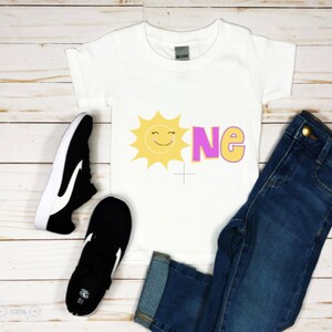 May include: White t-shirt with a yellow sun and pink "one" graphic. The shirt is paired with blue jeans and black sneakers.