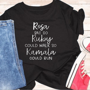 May include: Black t-shirt with white text that reads "Rosa sat so Ruby could walk so Kamala could run".