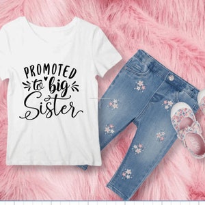 Può includere: Maglietta bianca con il testo "Promossa a sorella maggiore" in lettere nere. La maglietta è abbinata a jeans blu con ricami floreali e scarpe bianche con stampa floreale.