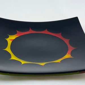 May include: A black square glass serving dish with a raised rim and a yellow, orange, and red sun design in the center.