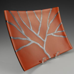 May include: A rectangular orange glass serving dish with a silver tree design. The dish is on a white stand.