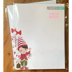 May include: A notepad with a festive elf design. The notepad is white with a cartoon elf wearing a red and white striped hat and red outfit. The notepad measures 4.1 x 5.45 inches and has the text "PHOTOCARDDESIGNS" and "NOTEPAD".
