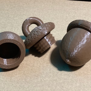 May include: Three brown acorn-shaped containers made of plastic. One container is open, showing the hollow interior and threaded rim. Another piece is a separate cap with a loop on top and threaded base. The third piece is a closed acorn.